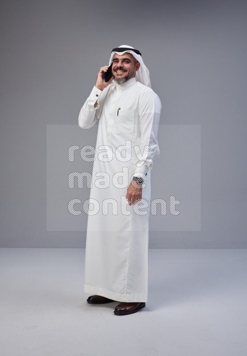Saudi man Wearing Thob and white Shomag standing talking on phone on Gray background