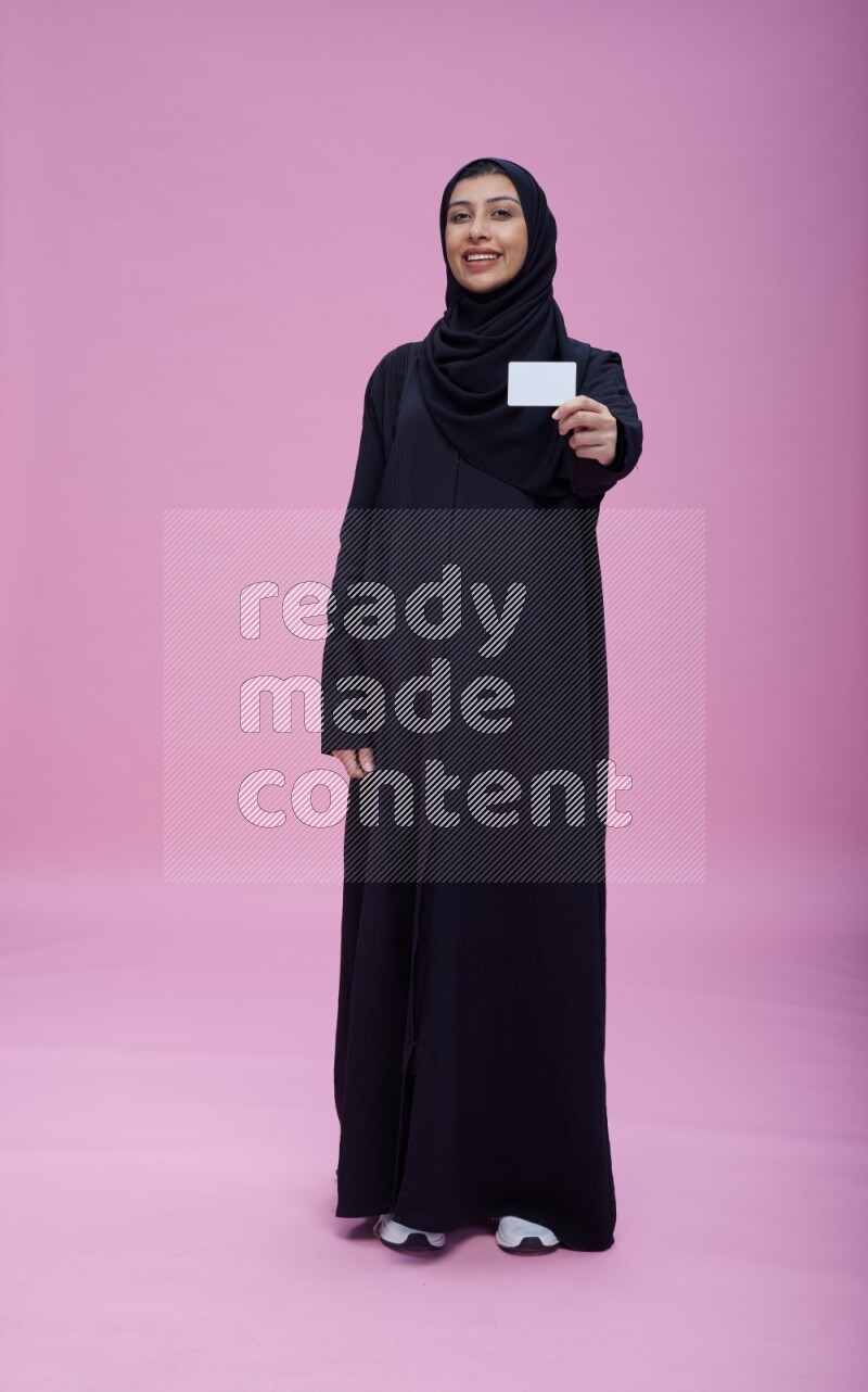 Saudi woman wearing Abaya standing holding ATM card on pink background
