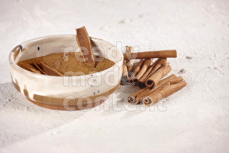 Ceramic bowl full of cinnamon powder with cinnamon sticks on the side on white background