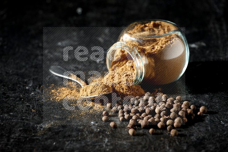 A flipped glass spice jar and metal spoon full of allspice powder and powder spilled out of it with whole balls on a textured black flooring