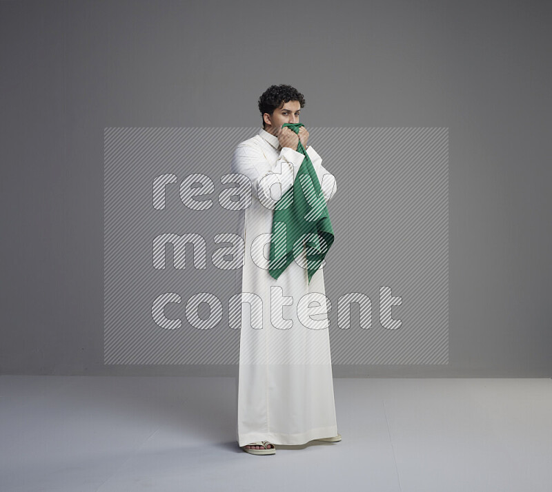 A Saudi man standing wearing thob kissing Saudi flag on gray background