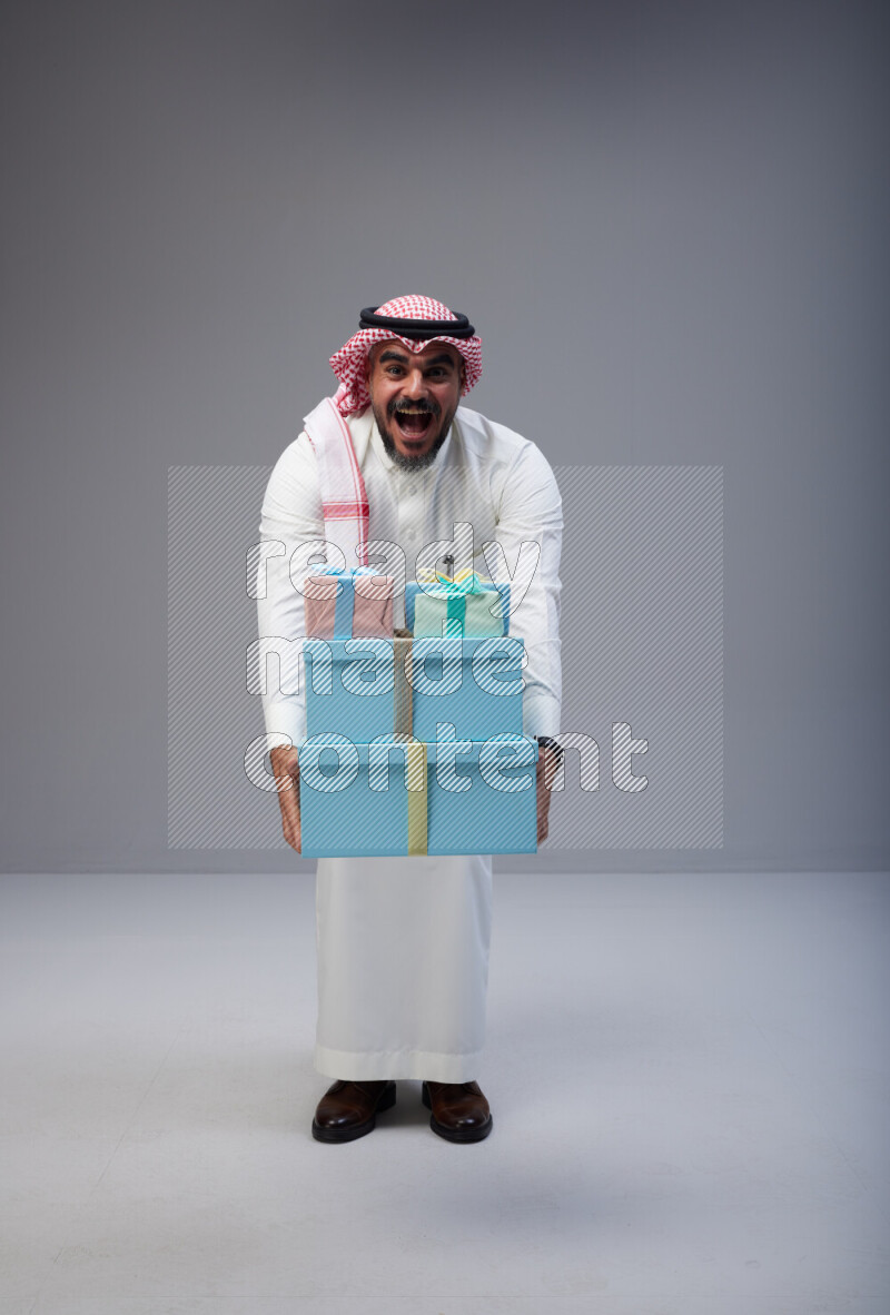 Saudi man Wearing Thob and red Shomag standing holding gift box on Gray background