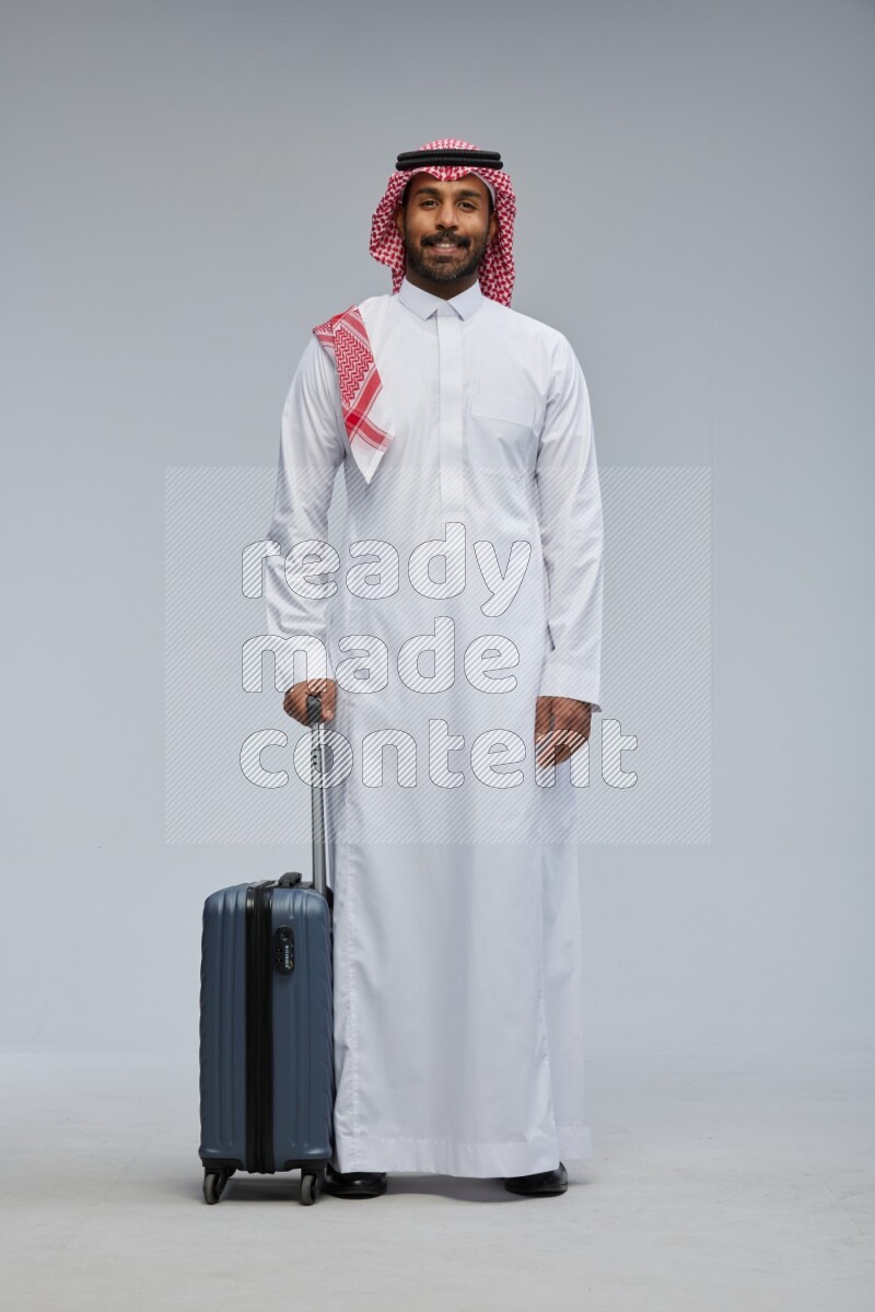 Saudi man wearing Thob and shomag standing holding Travel bag on Gray background