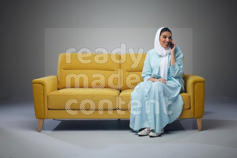 A Saudi woman wearing a light blue Abaya and a white head scarf sitting on a yellow sofa and talking in the phone while making different poses eye level on a grey background