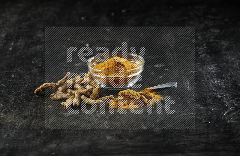 A glass bowl and a metal spoon full of turmeric powder and dried whole fingers next of them on textured black flooring