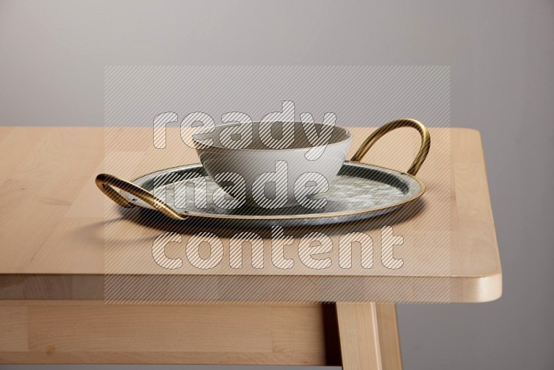 off white bowl with islamic pattern placed on a rounded stainless steel tray with golden handels on the edge of wooden table