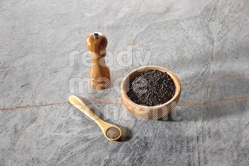 وعاء خشبي ممتلئ بالفلفل الأسود وملعقة خشبية ممتلئة بمسحوق الفلفل الأسود ومطحنة خشبية على خلفية رخام رمادية