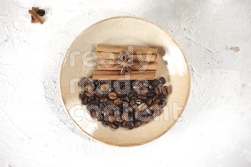 Beige plate full of coffee beans, cinnamon sticks and star anise with a coffee grinder, coffee beans, cinnamon pieces and cardamom next of it on white background