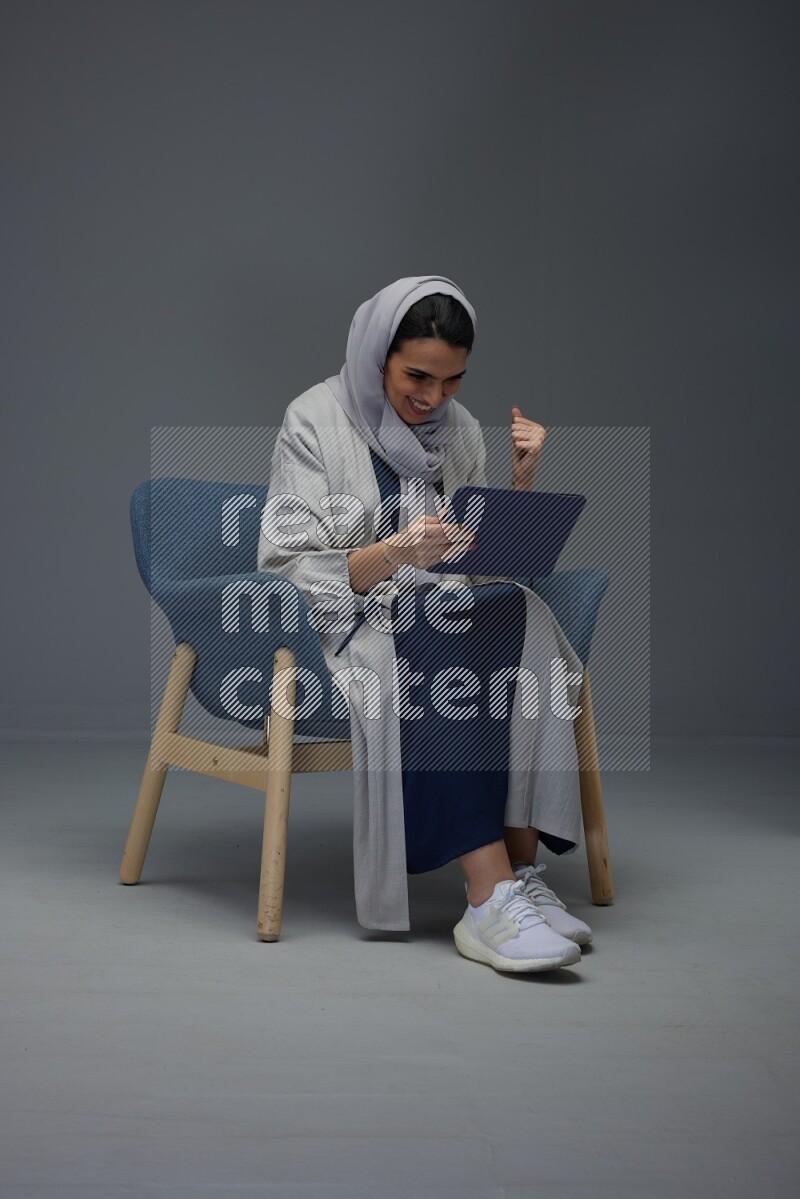 A Saudi female wearing a light gray Abaya and head scarf sitting on a dark grey chair while holding her iPad on a grey background