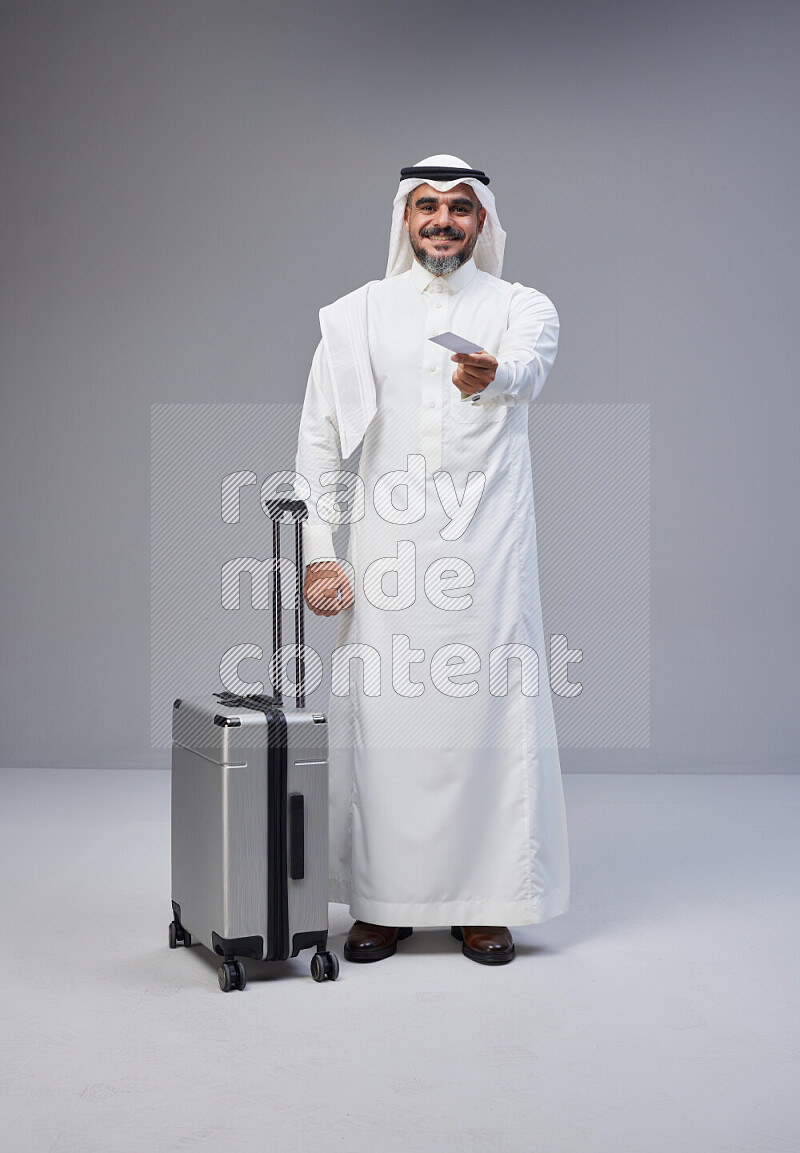 Saudi man wearing Thob and white Shomag standing holding Travel bag and ATM card on Gray background