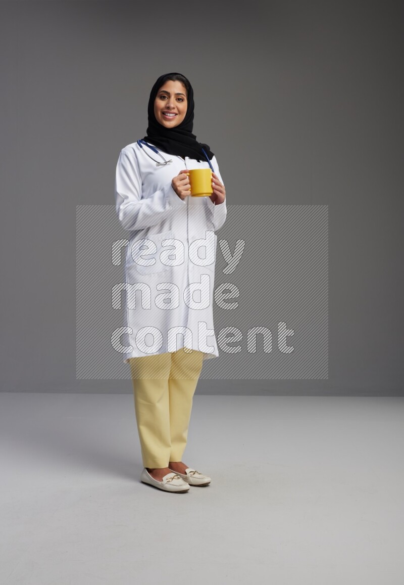 Saudi woman wearing lab coat with stethoscope standing holding mug on Gray background