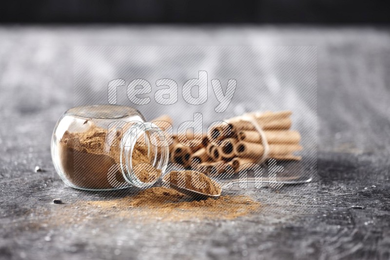 Herbal glass jar full cinnamon powder flipped and a metal spoon full of powder, cinnamon sticks stacked and bounded in the back on textured black background in different angles