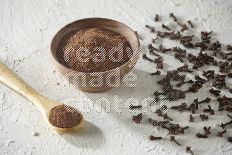 A wooden bowl and wooden spoon full of cloves powder with cloves spread on textured white flooring