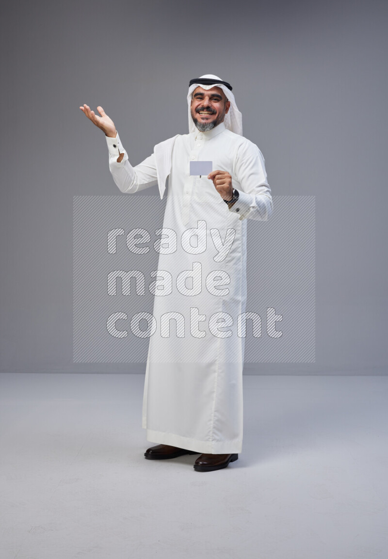 Saudi man Wearing Thob and white Shomag standing holding ATM card on Gray background