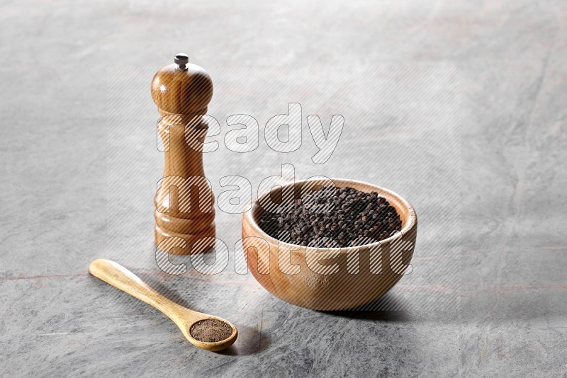 A wooden bowl full of black pepper and a wooden spoon full of black pepper powder and a wooden grinder on a marble flooring