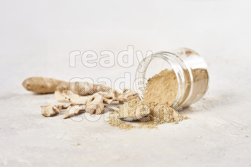 A glass jar full of ground ginger powder flipped with some spilling powder on white background