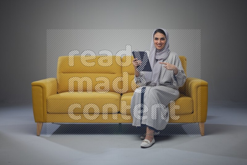 A Saudi woman wearing a light gray Abaya and head scarf sitting on a yellow sofa and holding her tablet while pointing to it eye level on a grey background