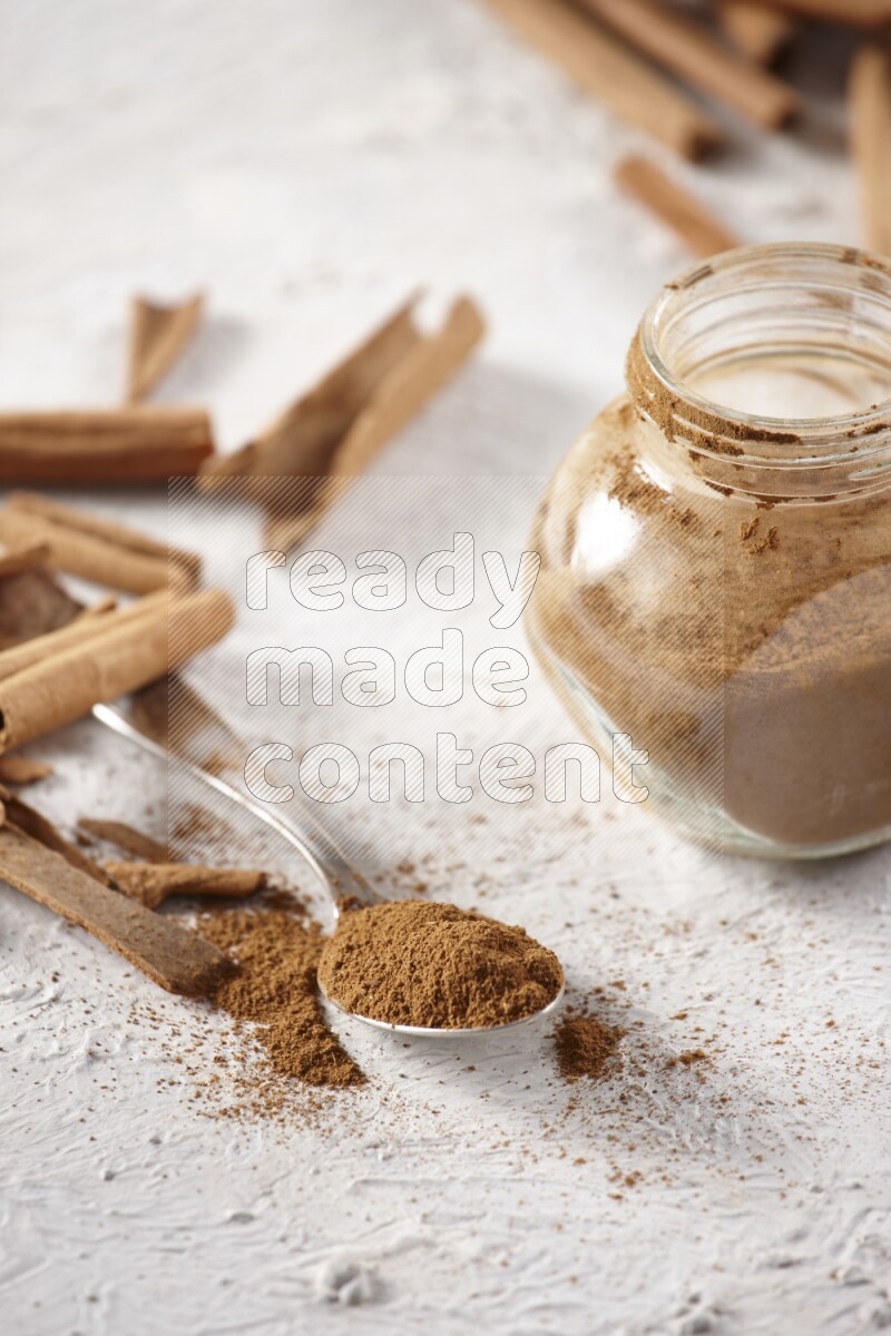 Herbal glass jar full cinnamon powder and a metal spoon surrounded by cinnamon sticks on a white background