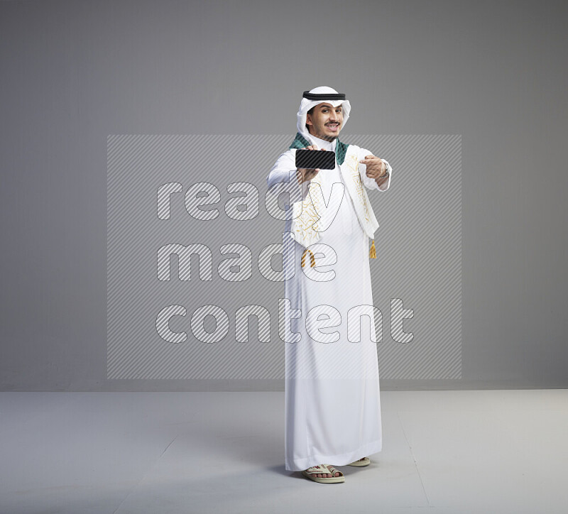 A Saudi man standing wearing thob and white shomag with flag scarf on his neak showing phone on gray background