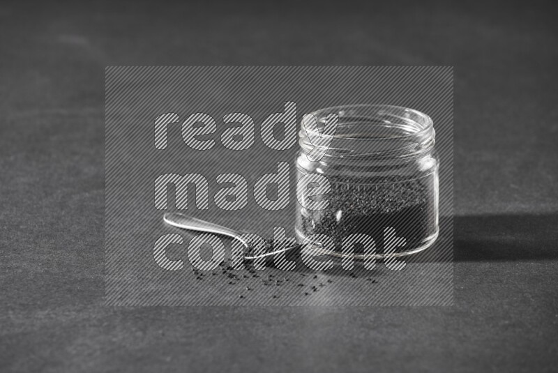 A glass jar full of black seeds with a metal spoon full of the seeds on a black flooring