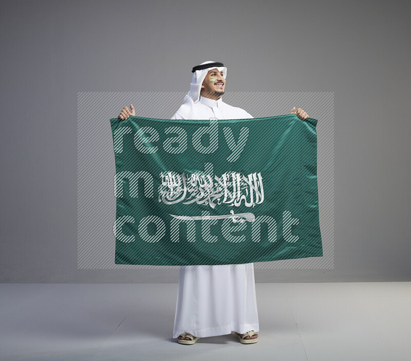 A Saudi man standing wearing thob and white shomag with face painting holding big Saudi flag on gray background