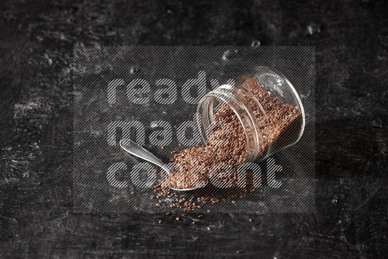 A glass jar full of flax flipped and seeds spreaded out with a metal spoon full of the seeds on a textured black flooring in different angles
