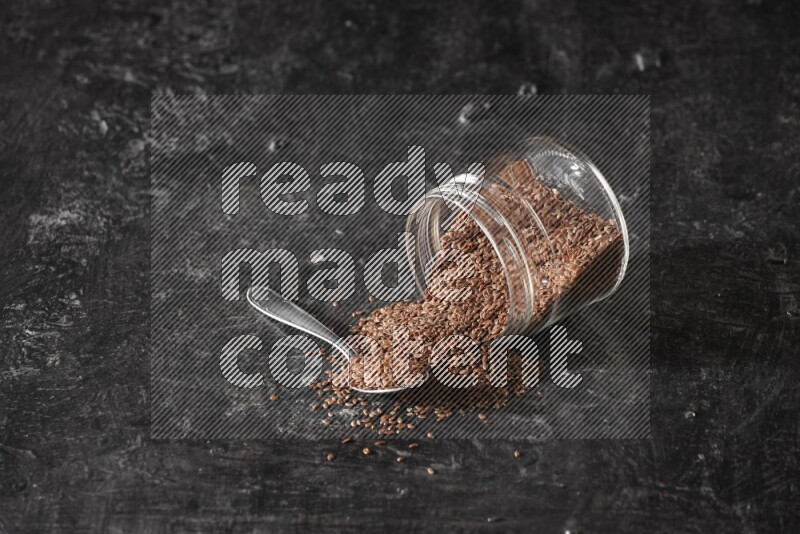 A glass jar full of flaxseeds flipped and seeds spread out with a metal spoon full of the seeds on a textured black flooring