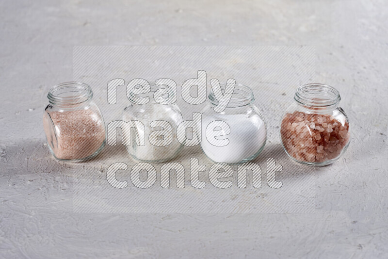 4 glass jars filled with table salt, coarse sea salt, fine himalayan salt and coarse himalayan salt on white background