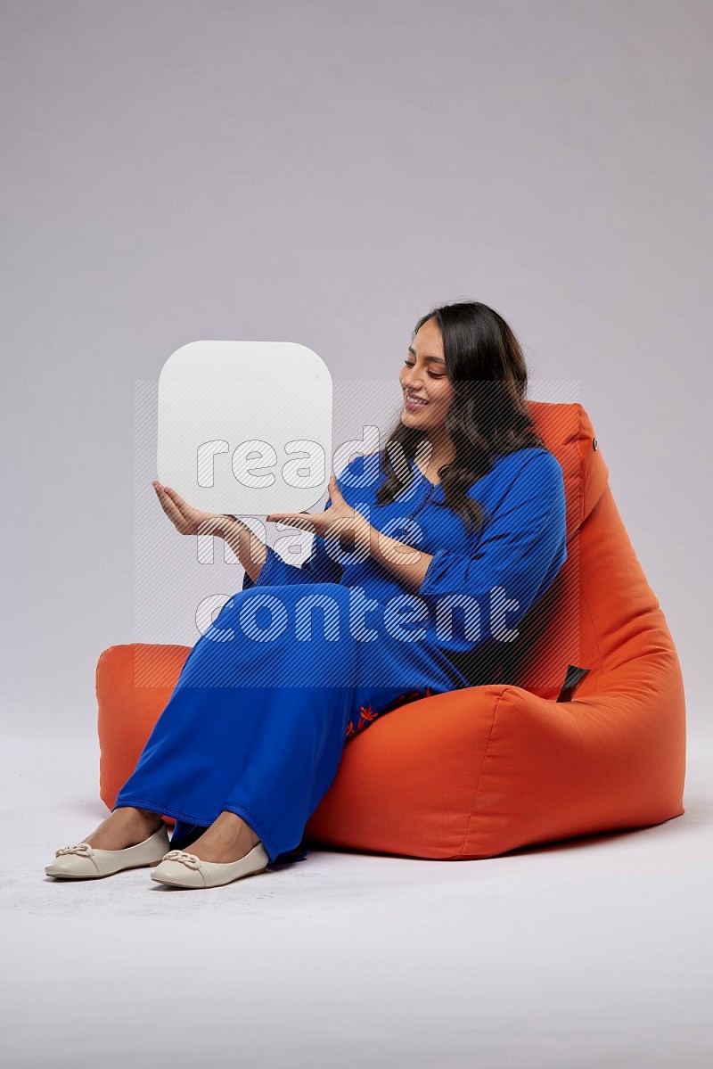 A Woman sitting on an orange beanbag wearing Jalabeya holding a social media sign