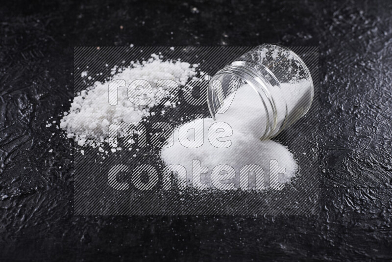 A glass jar full of table salt with some sea salt crystals beside it on a black background