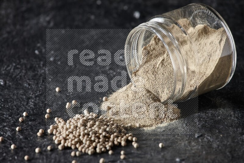 A flipped glass jar full of white pepper powder with spilled powder and pepper beads on textured black flooring