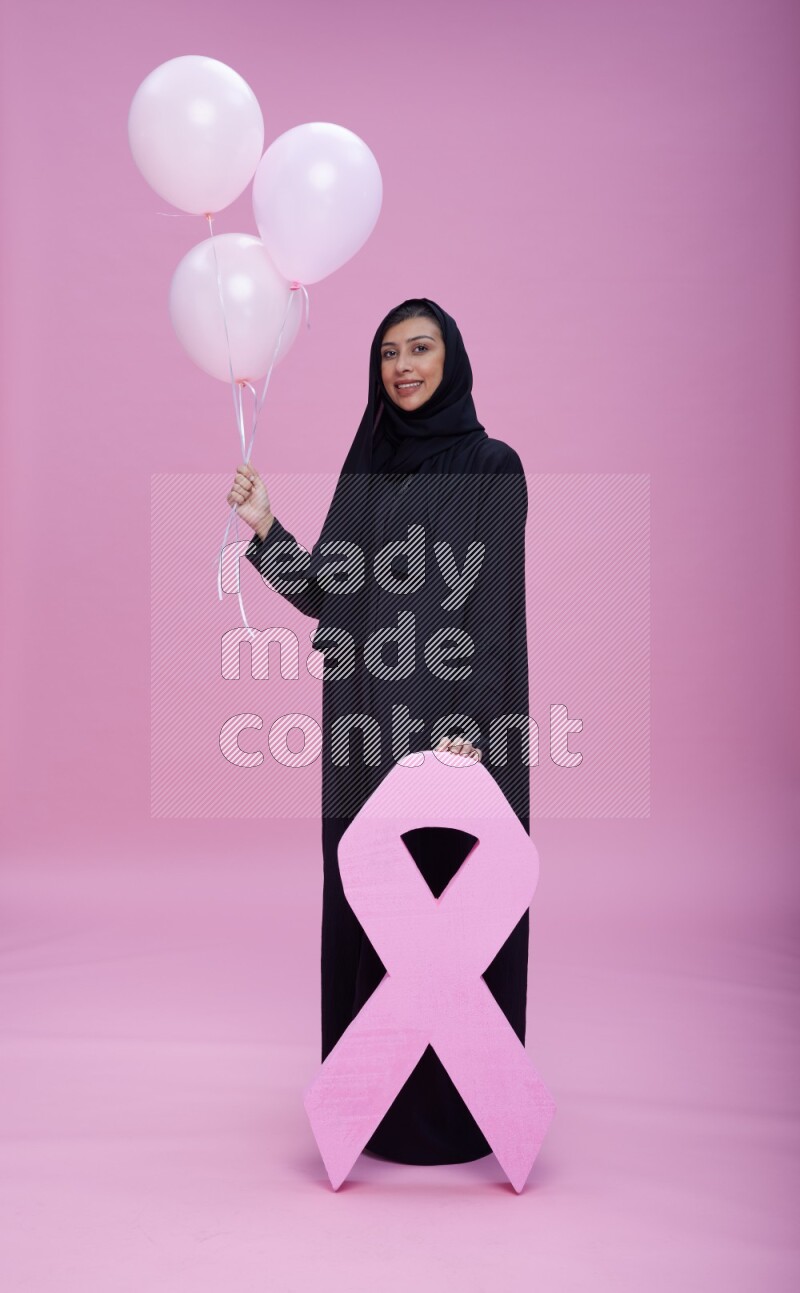 Saudi woman wearing pink ribbon on Abaya standing holding balloons and awareness sign on pink background