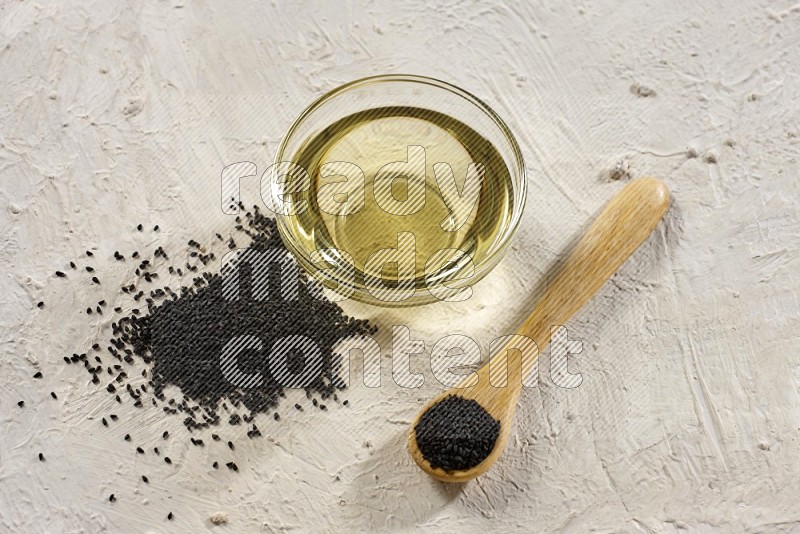 A glass bowl full of black seeds oil and wooden spoon full of black seeds with seeds spread on a textured white flooring