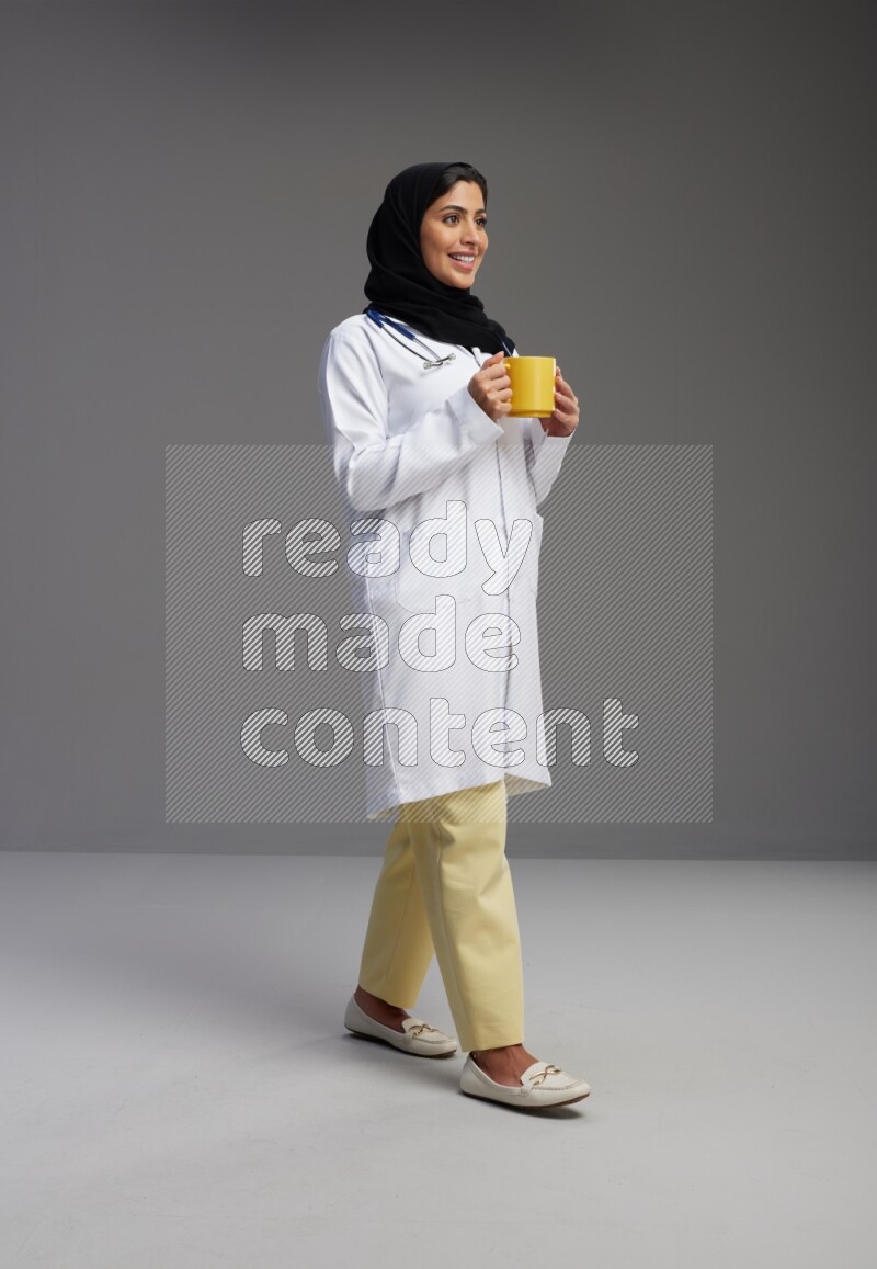 Saudi woman wearing lab coat with stethoscope standing holding mug on Gray background