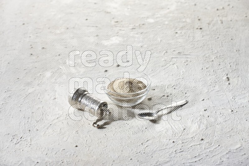 A glass bowl full of white pepper powder with white pepper beads and a metal grinder on textured white flooring