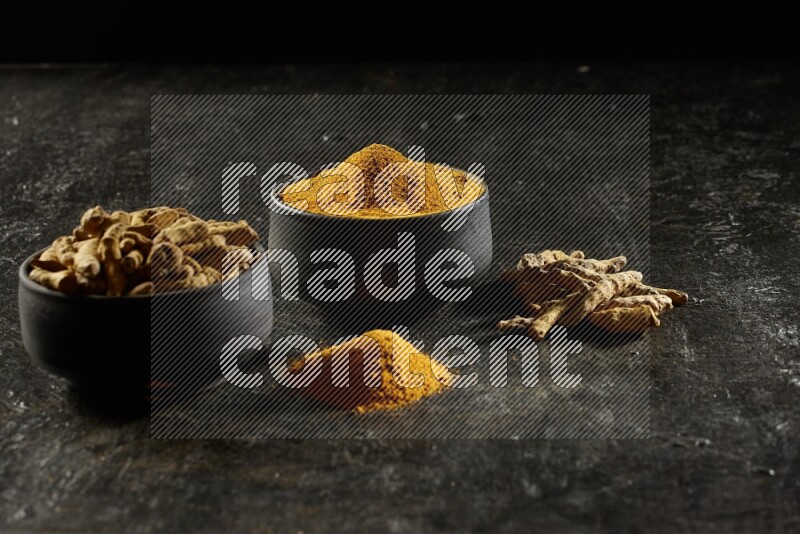 2 pottery black bowls, one full of turmeric powder and the other full of dried turmeric whole fingers on a textured black flooring