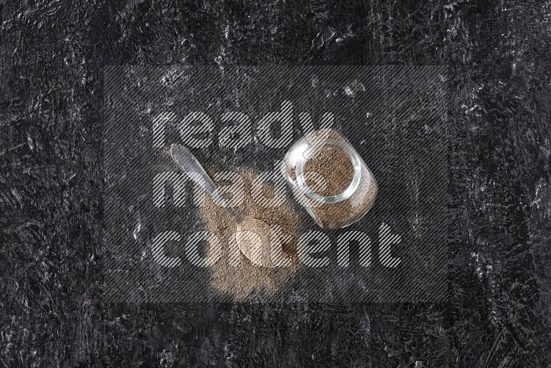 بودرة الفلفل الأسود في وعاء زجاجي مع ملعقة معدنية مليئة بالبودرة علي خلفية سوداء