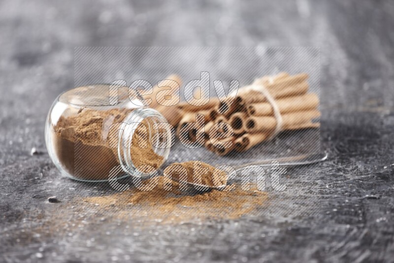 Herbal glass jar full cinnamon powder flipped and a metal spoon full of powder, cinnamon sticks stacked and bounded in the back on textured black background in different angles
