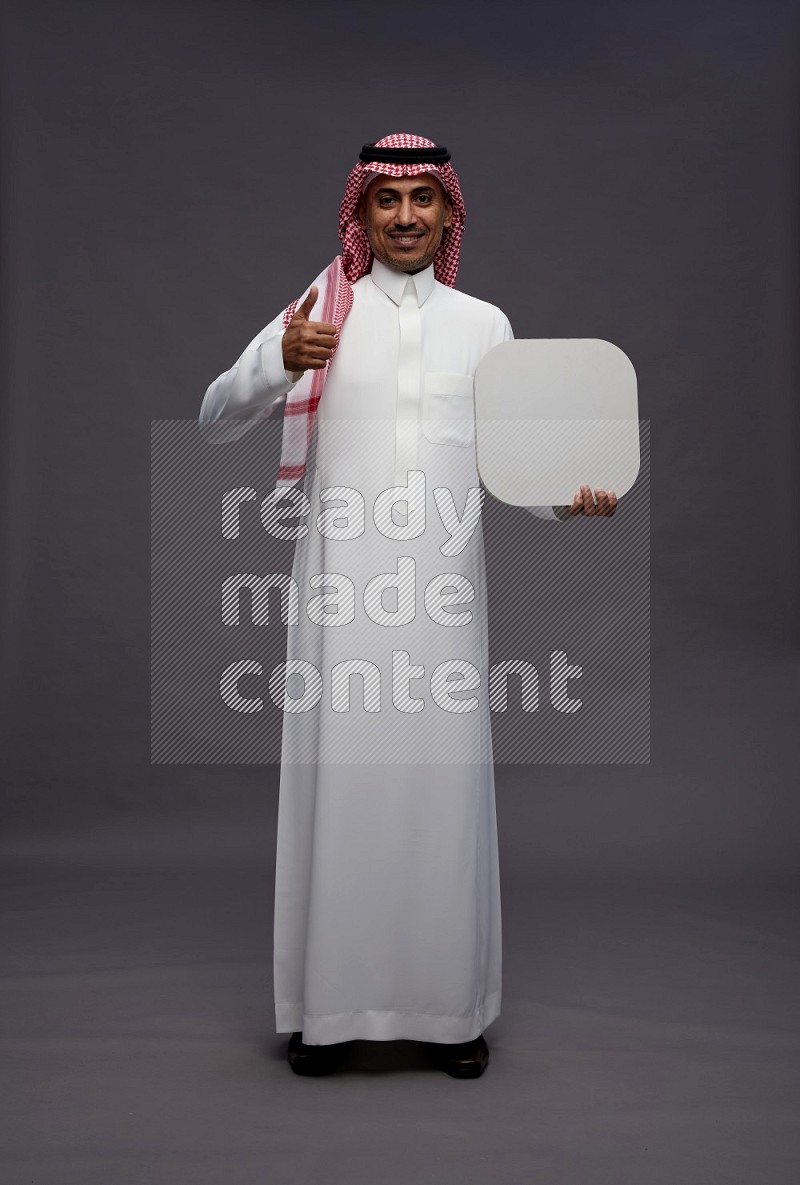 Saudi man wearing thob and shomag standing holding social media sign on gray background