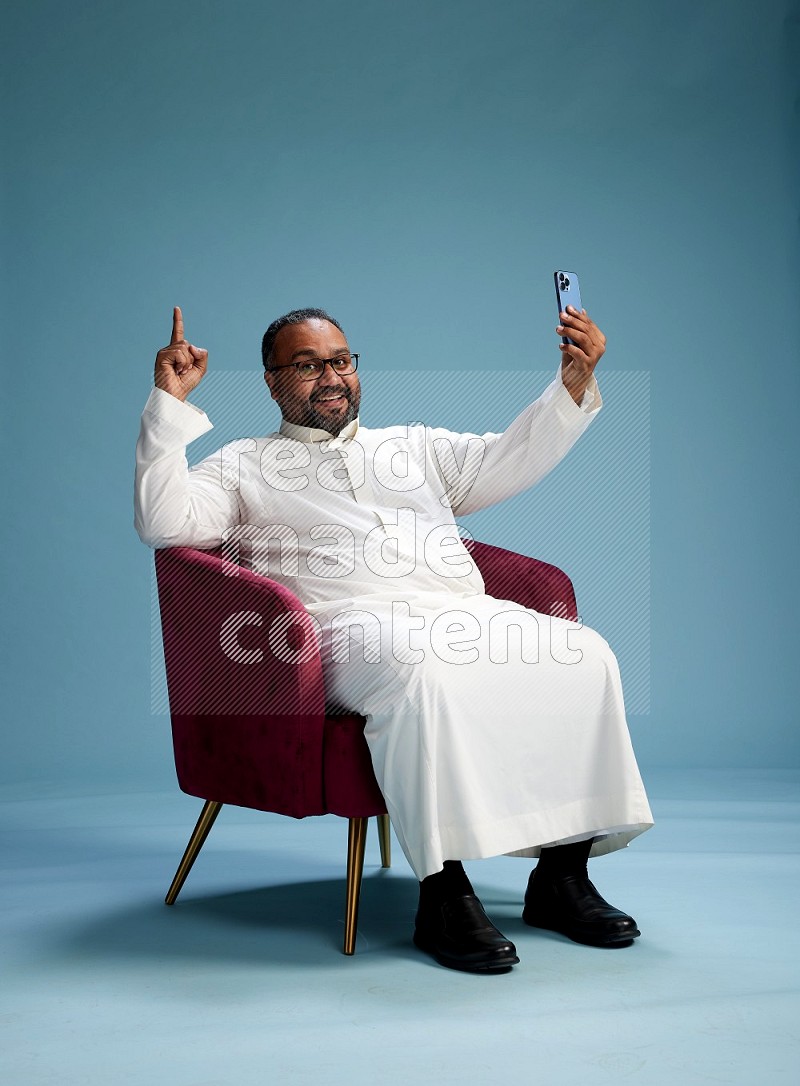 Saudi Man without shimag sitting on chair taking selfie on blue background
