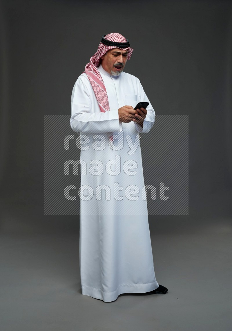 Saudi man with shomag Standing texting on phone on gray background