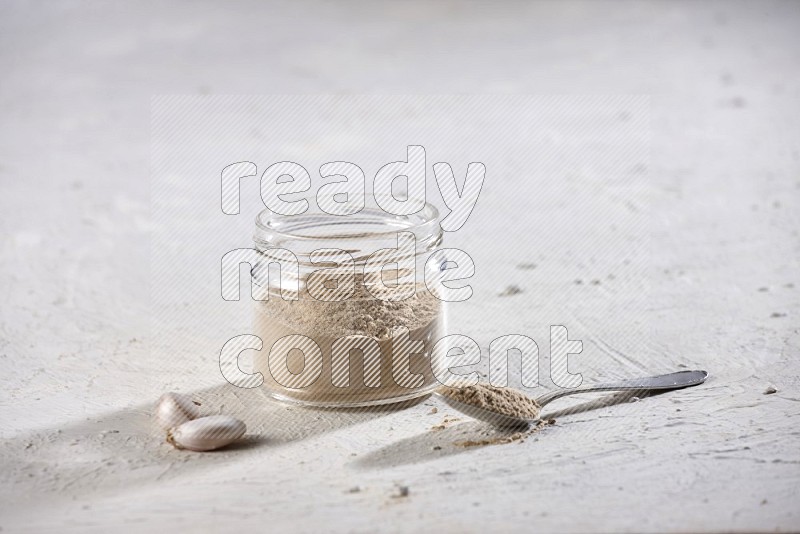 A glass jar full of garlic powder with metal spoon full of the powder on a textured white flooring in different angles