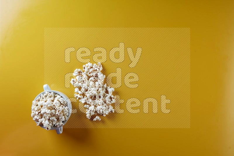 A white ceramic bowl full of popcorn with popcorn beside it on a yellow background in a top view shot