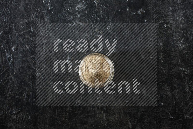 A glass jar full of cardamom powder on textured black flooring