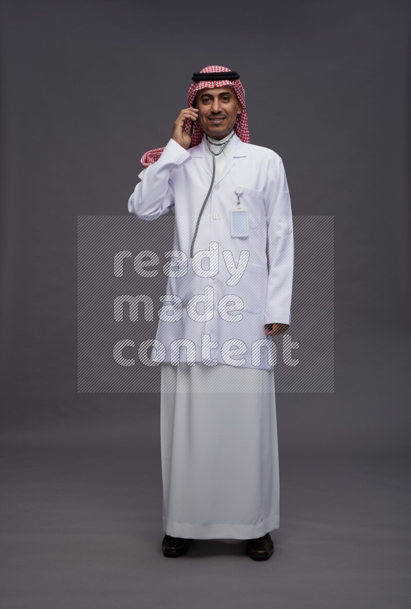 Saudi man wearing thob with lab coat and shomag with pocket employee badge standing talking on phone on gray background