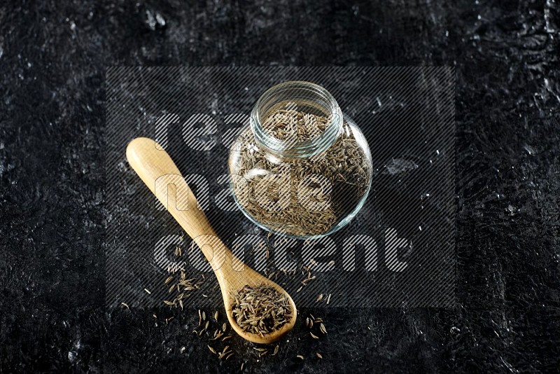 A glass spice jar and a wooden spoon full of cumin seeds on a textured black flooring