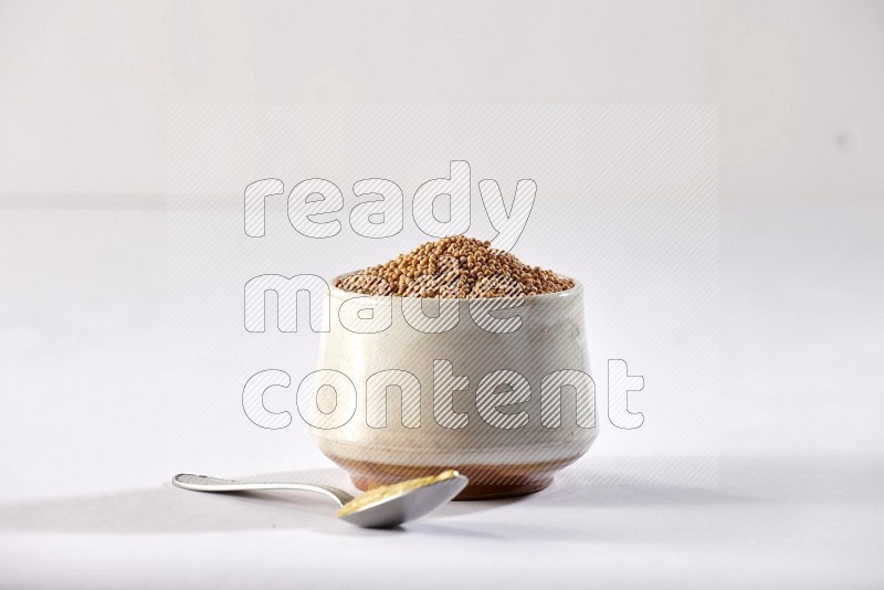 A beige pottery bowl full of mustard seeds and a metal spoon full of mustard paste on white flooring