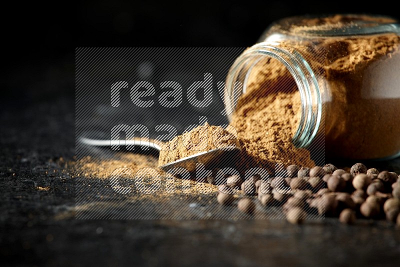 A flipped glass spice jar and metal spoon full of allspice powder and powder spilled out of it with whole balls on a textured black flooring