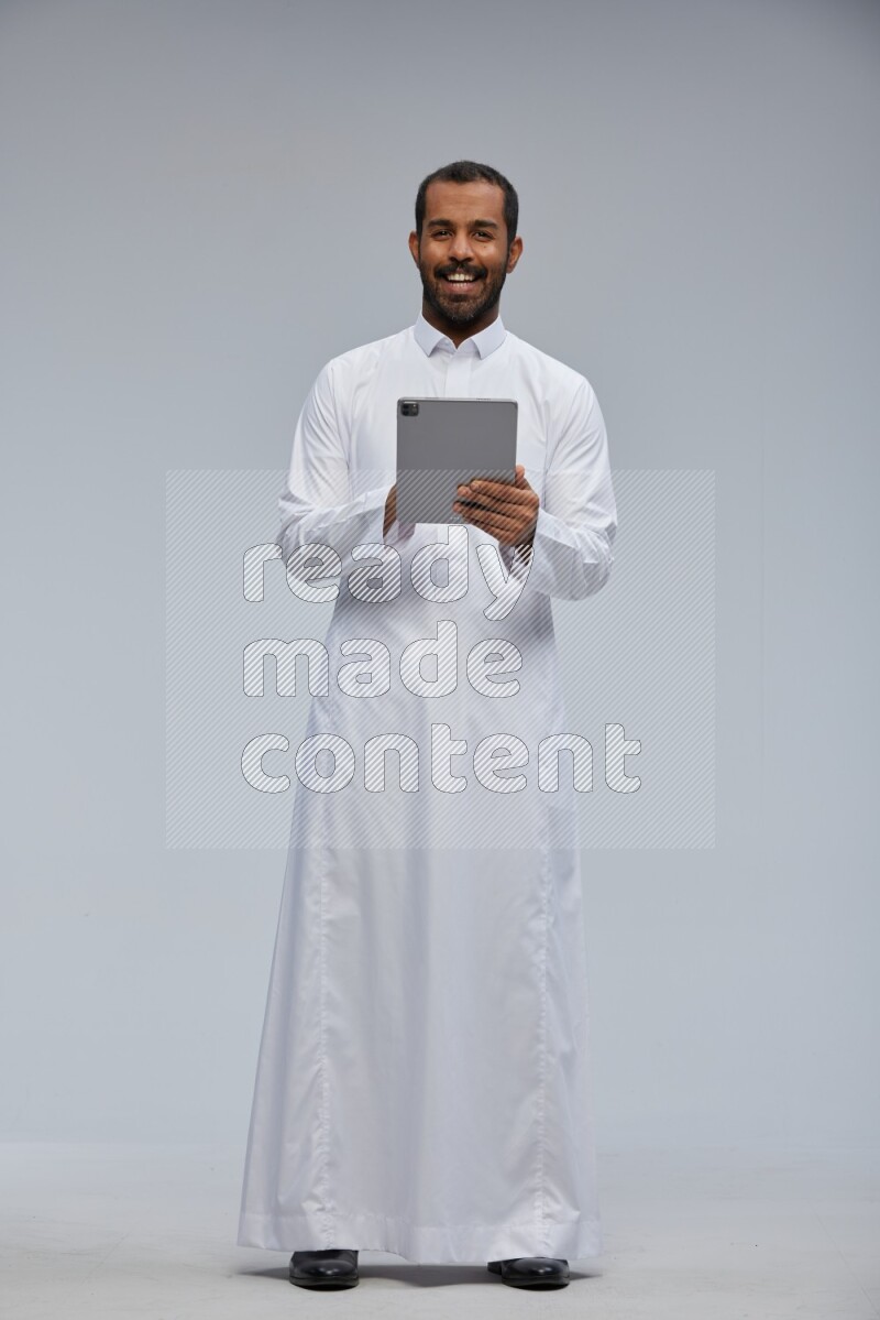 Saudi man wearing Thob standing working on tablet on Gray background