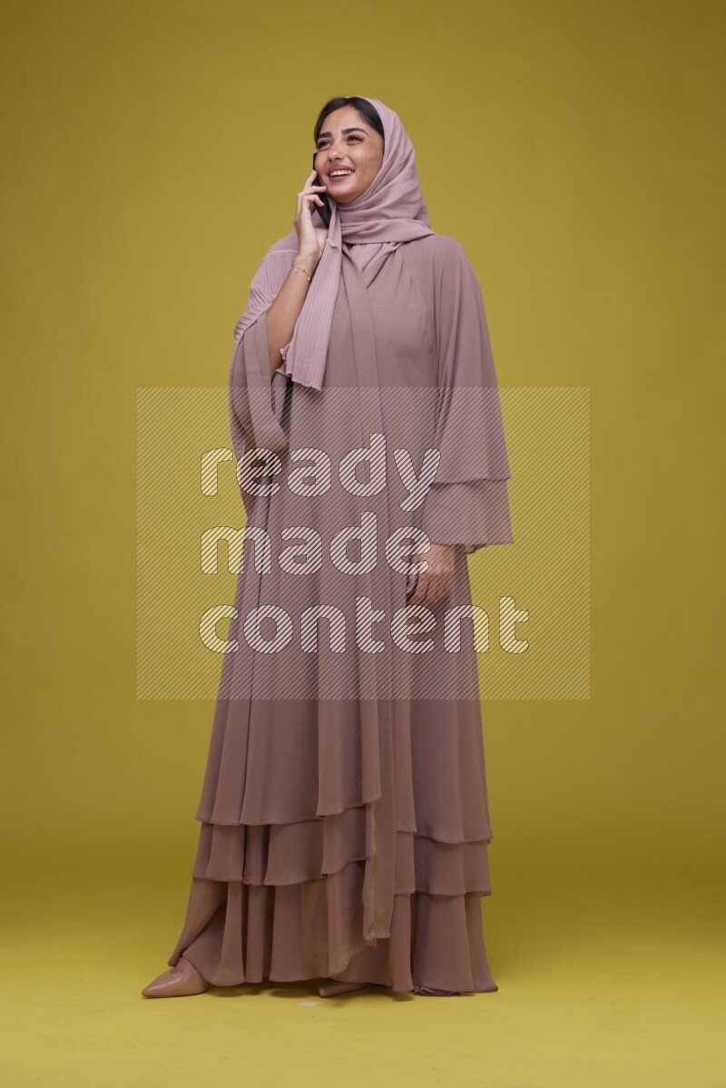 A woman having a call on a Yellow Background wearing Brown Abaya with Hijab
